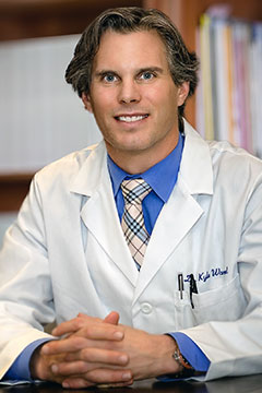 headshot of man in white lab coat grinning with hands interlocked seated at desk